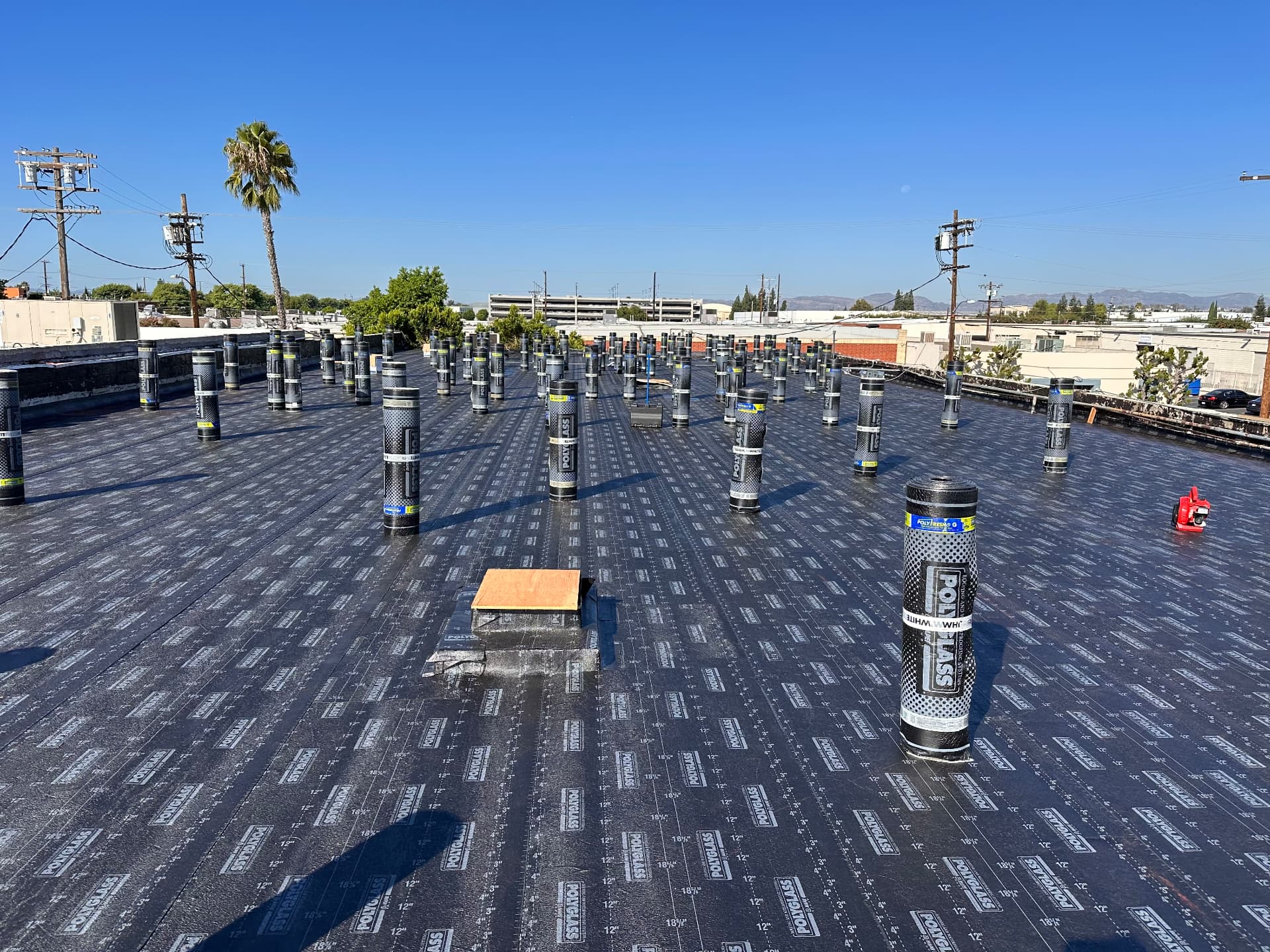 Large flat roof with base sheet applied and cap sheet rolls standing upright across the deck